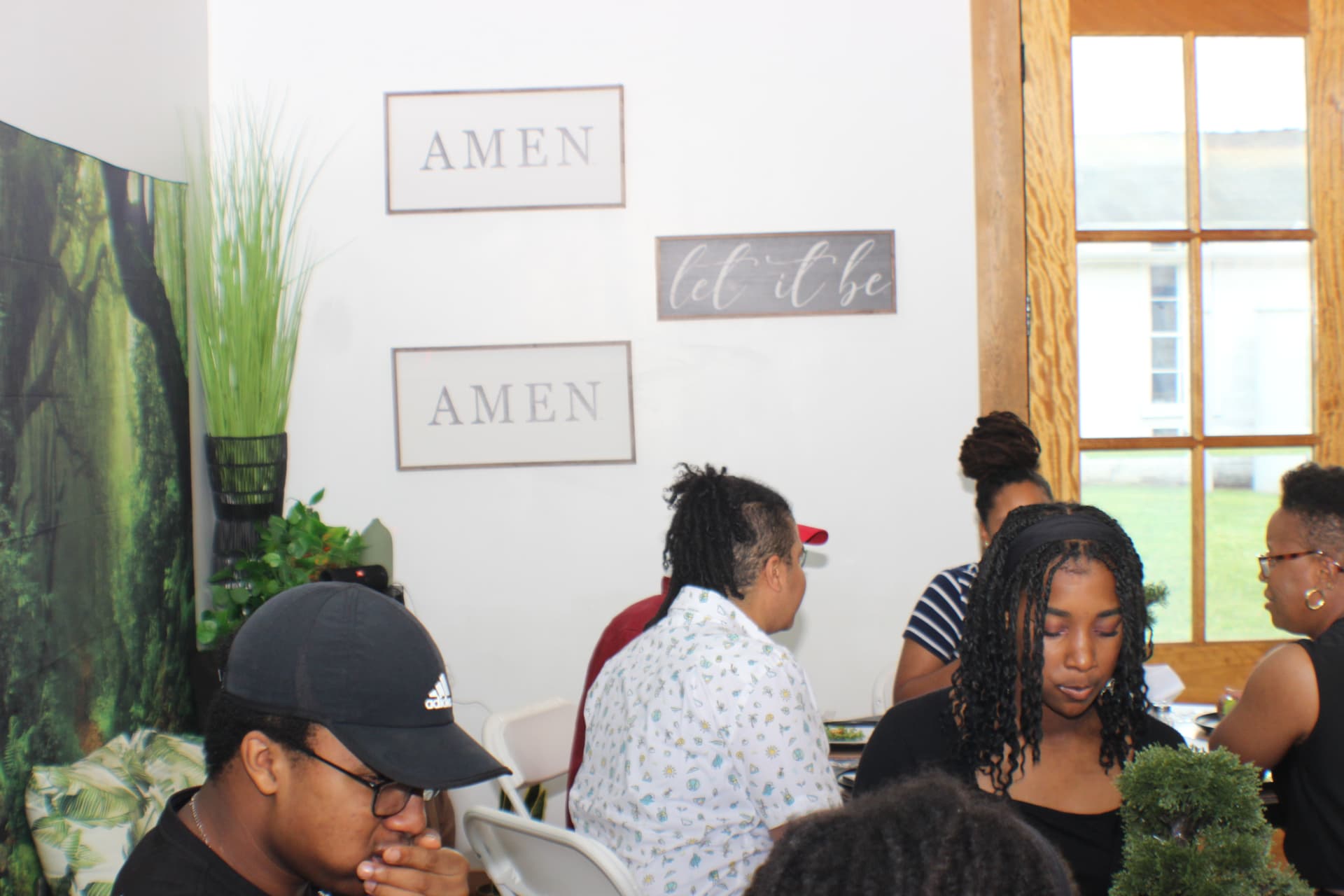 Community members seated together around a table indoors, beneath AMEN and let it be wall signs